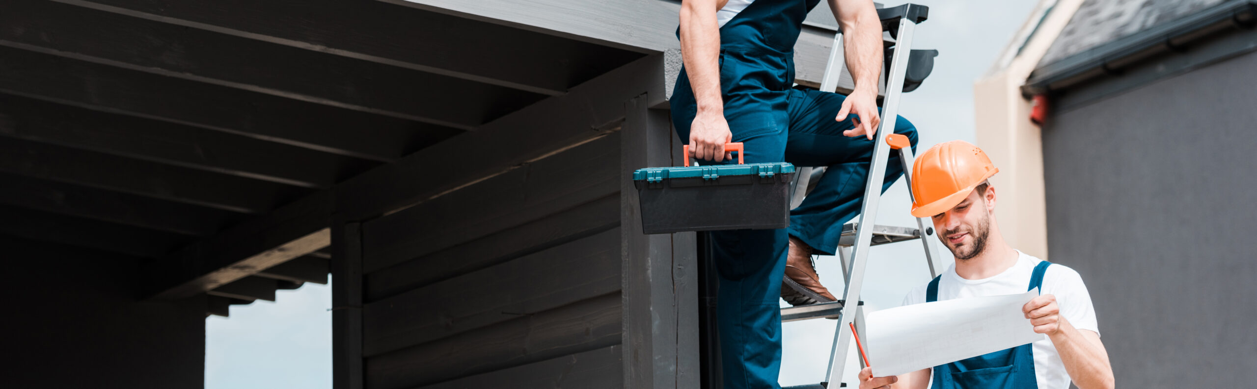 Two men working on a roof, one on a ladder, one holding plans.