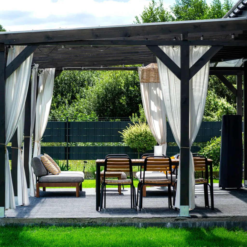 Outdoor patio with wooden pergola, white curtains, and dining set on a sunny day.