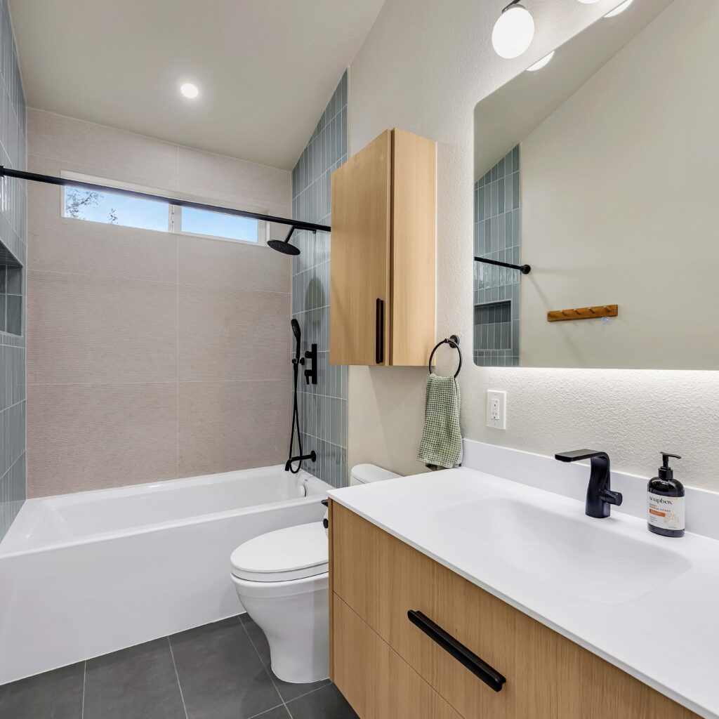 Modern bathroom with light wood vanity, white tub, and blue-gray tiles.