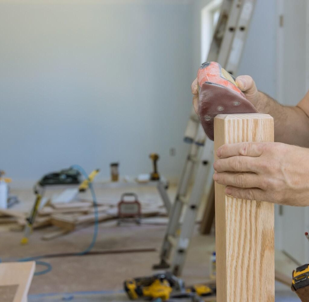 Person sanding a wooden newel post in a house under construction.