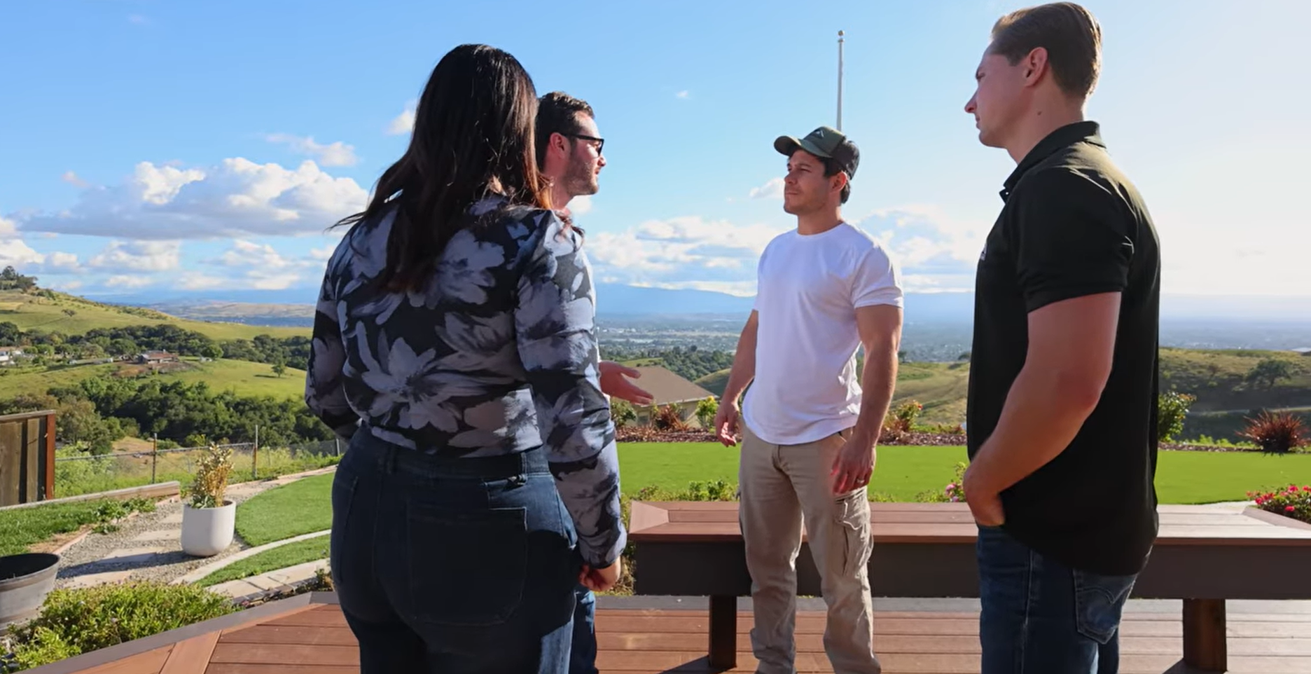 Four people standing on a wooden deck, talking, with a scenic outdoor view.